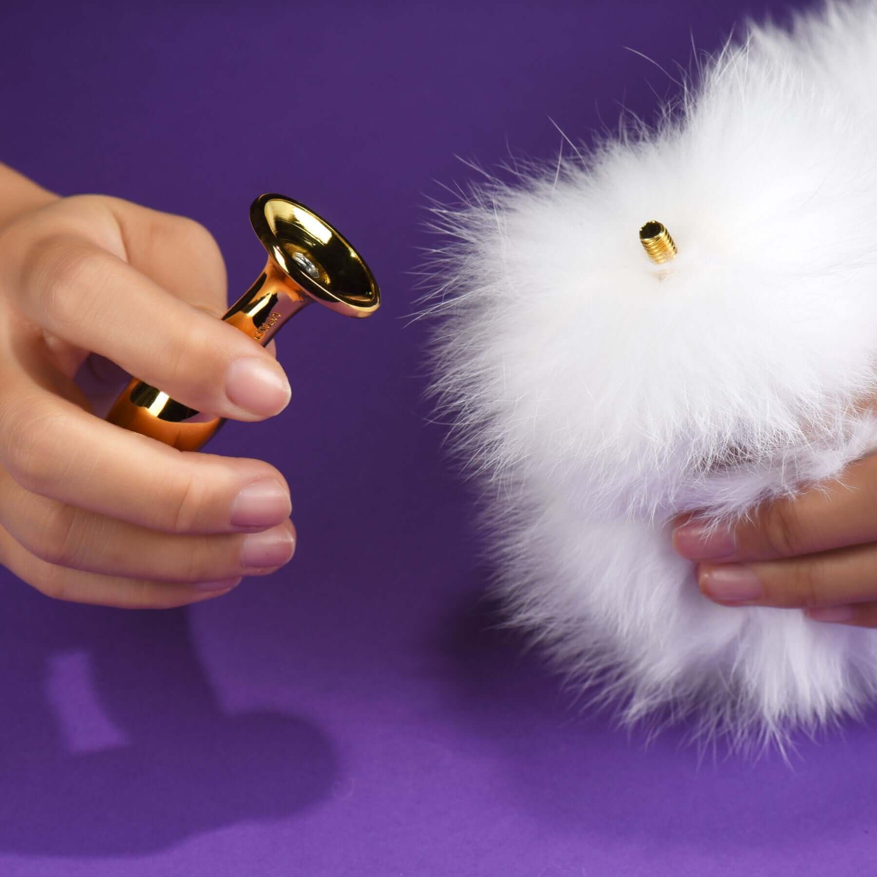 Hands holding a 24K gold-plated metal anal plug next to a fluffy white fox fur tail on a purple background.