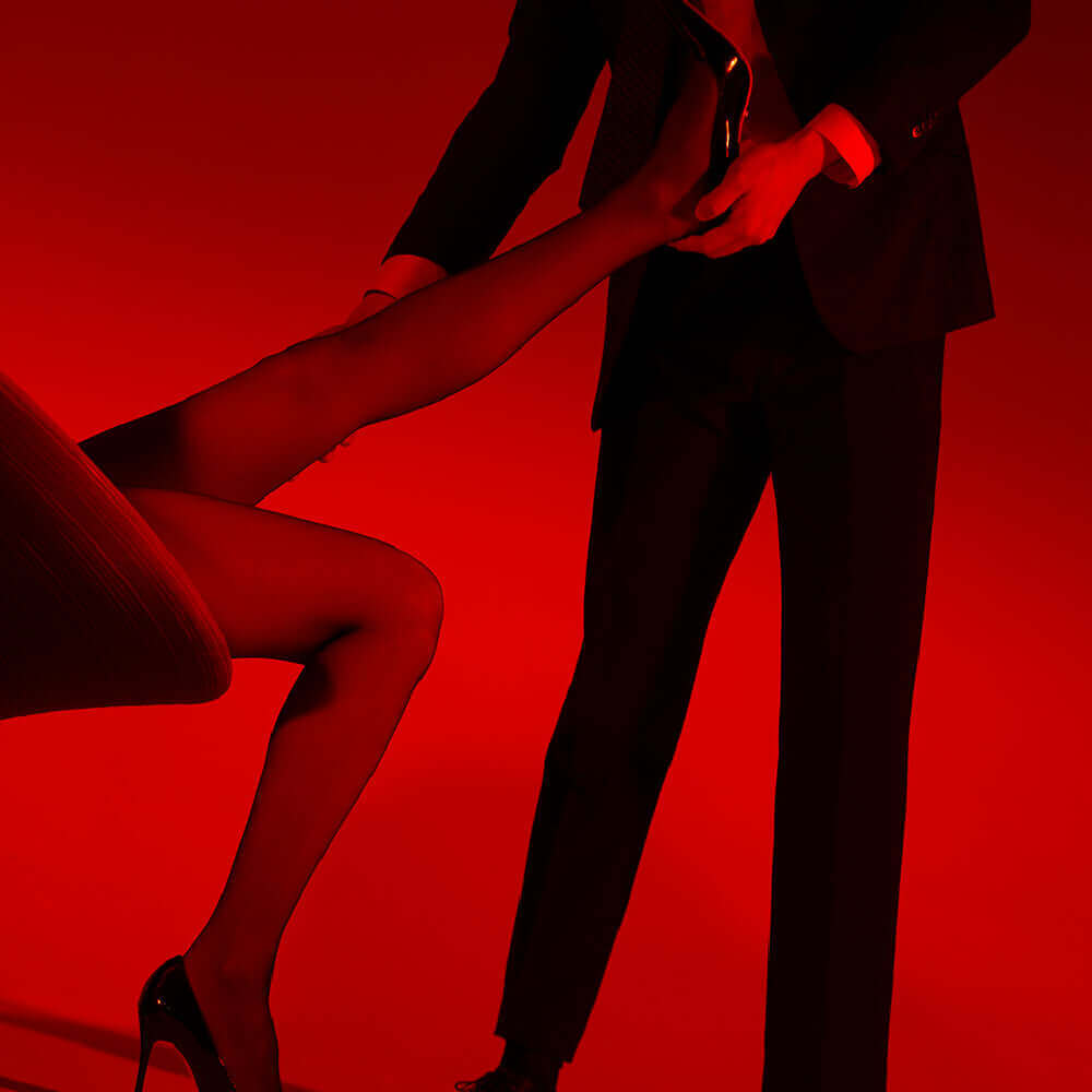 Elegant close-up of a man lifting a woman's leg, showcasing the UPKO 5D Tear-off Tights against a dramatic red backdrop.