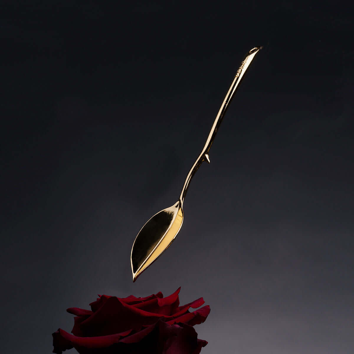 Elegant gold candle wax scraper hovering above a red rose on a dark background.