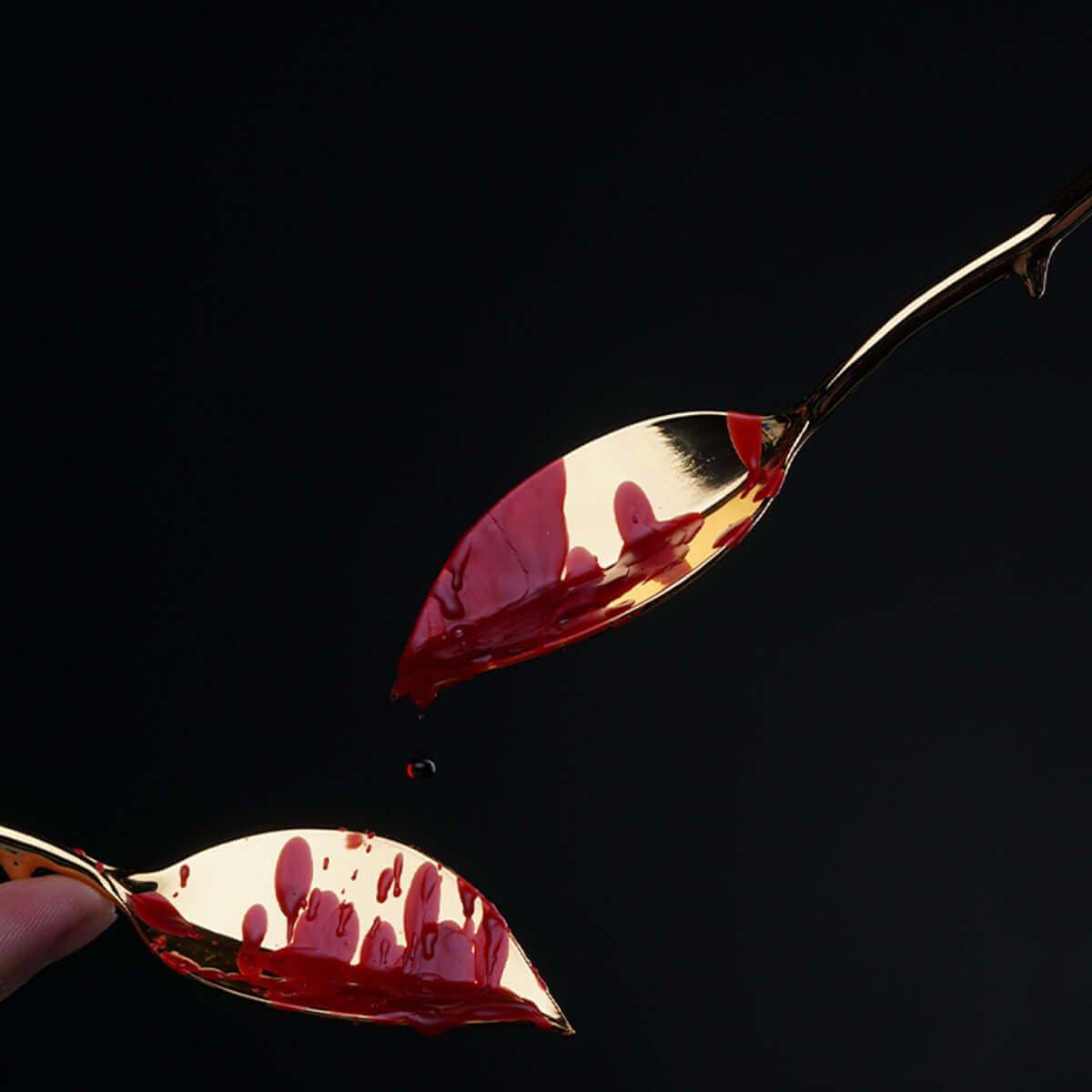 Two elegant golden spoons with red liquid dripping against a dark background.