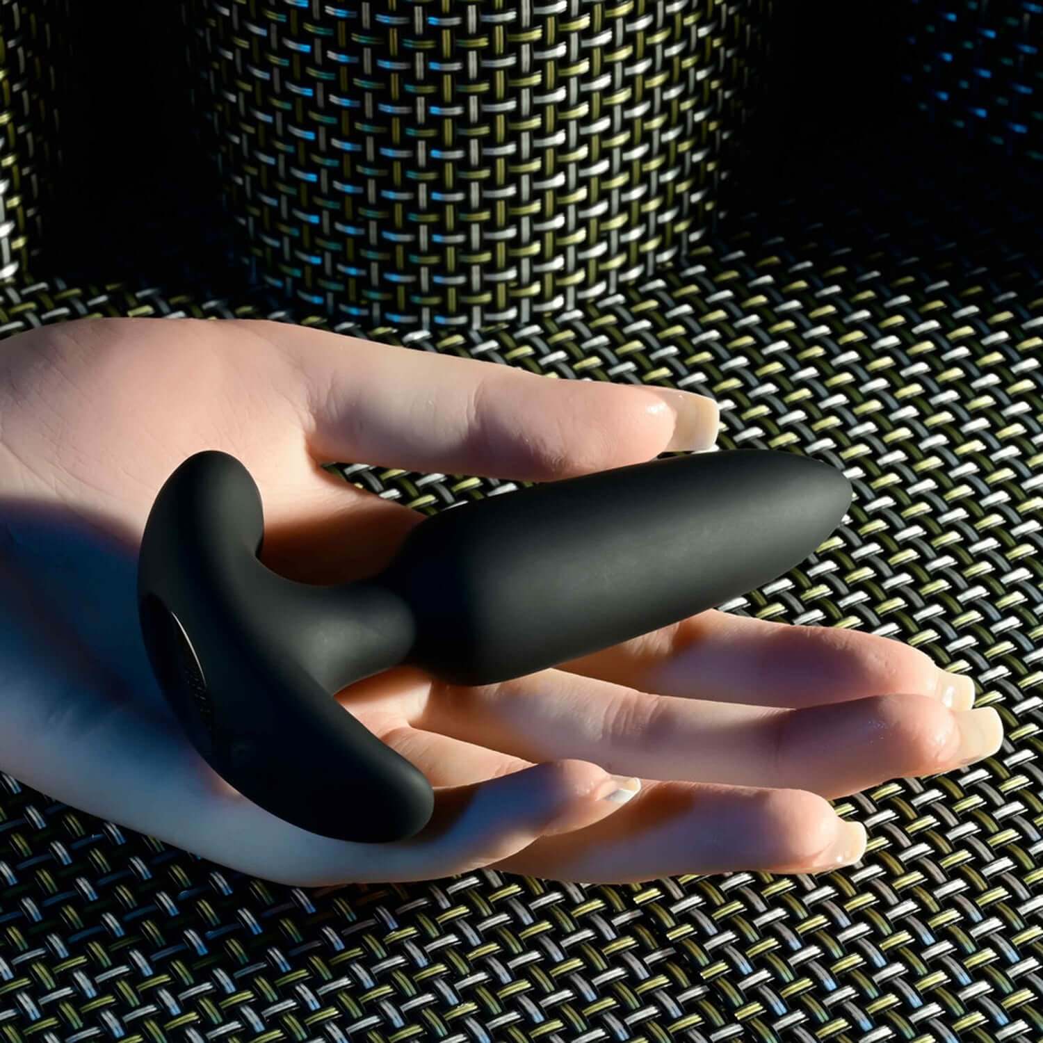 Black remote control anal plug held in a hand, showcasing its sleek design and tapered tip against a textured background.