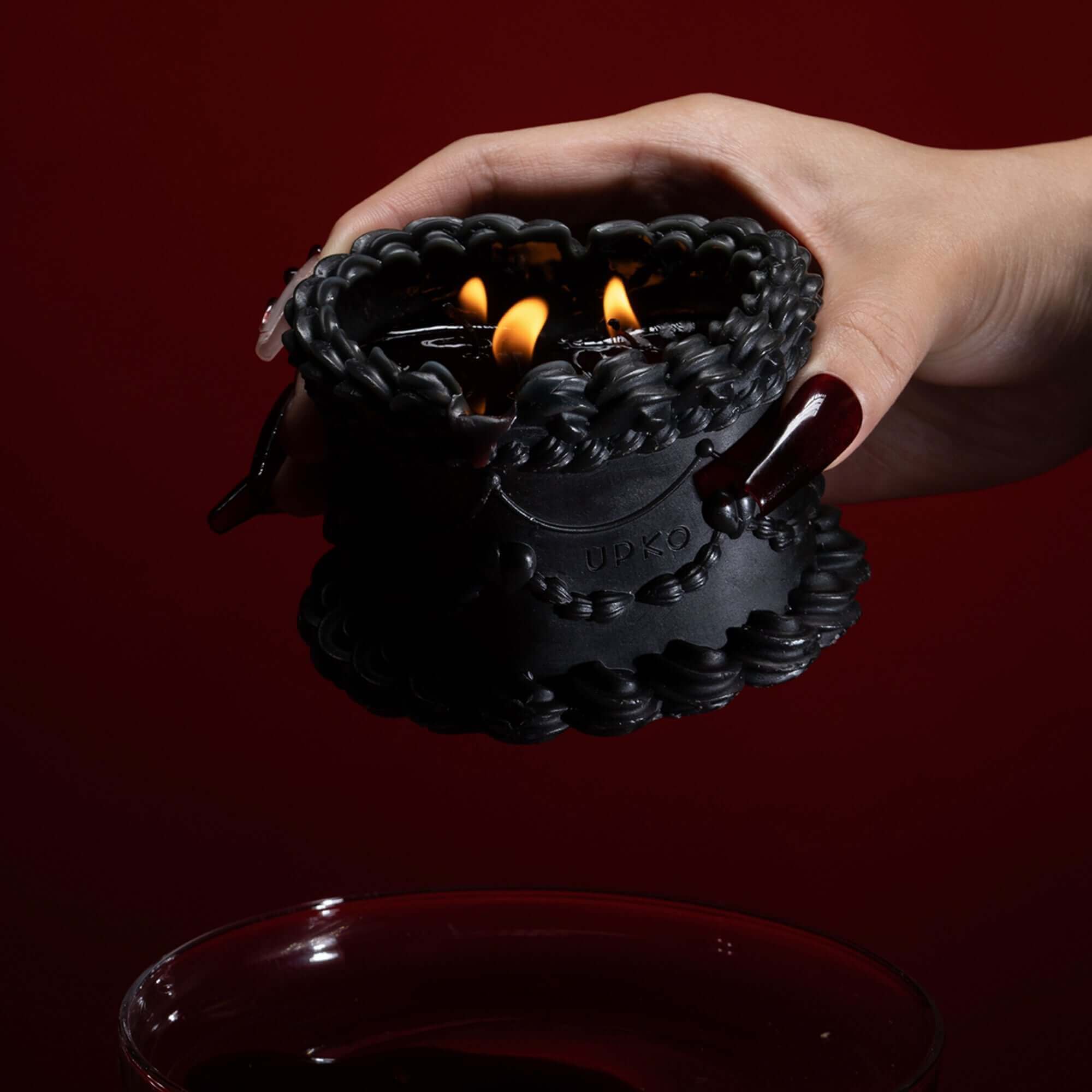 Hand holding a black low-temperature wax candle with lit flames, set against a deep red background.