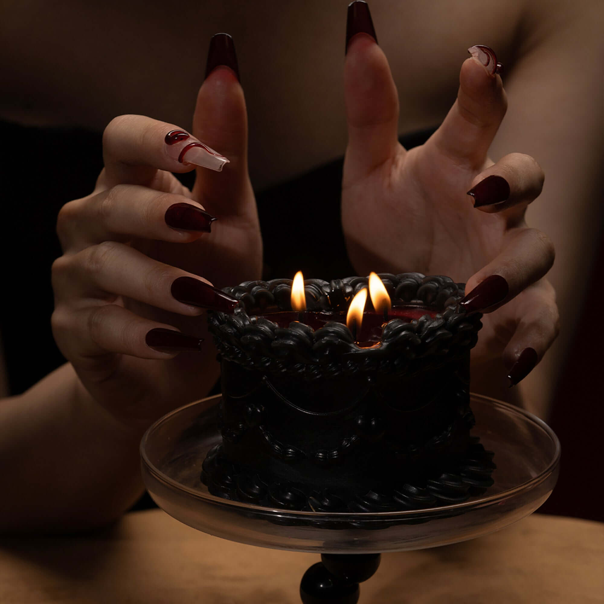 Hands with long red nails gently hold a black wax candle with flames, showcasing its elegant cake design.