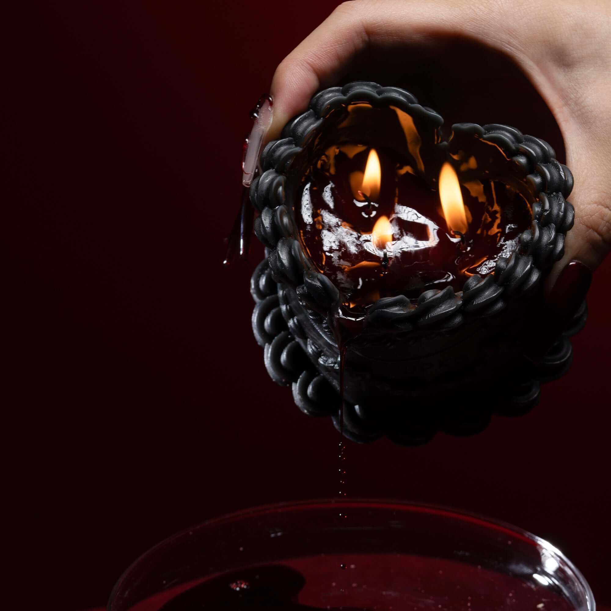 Heart-shaped wax candle with two flames, held above a bowl, showcasing its stylish red-and-black design.