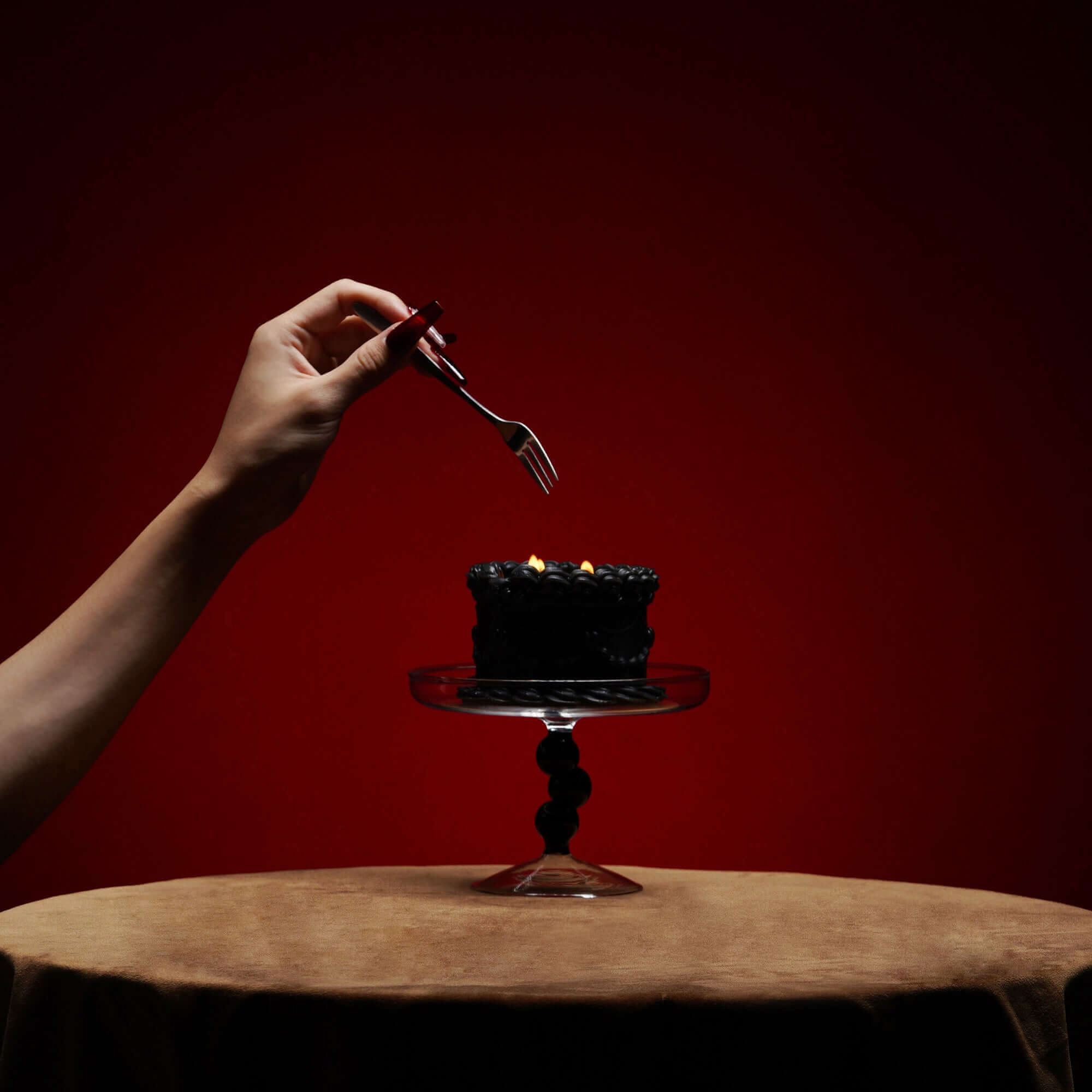 Hand holding a fork above a black cake on a glass stand with a red background, showcasing a stylish dessert presentation.