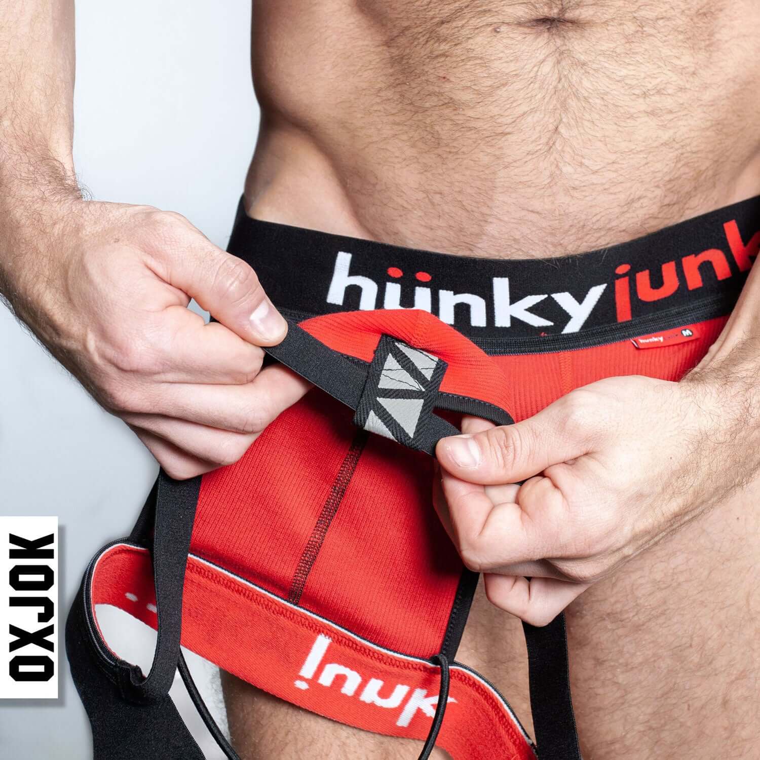 Man adjusting Hunkyjunk red jockstrap with comfy-pouch and oxjok butt-straps for no-dig comfort and bulge-lifting fit.