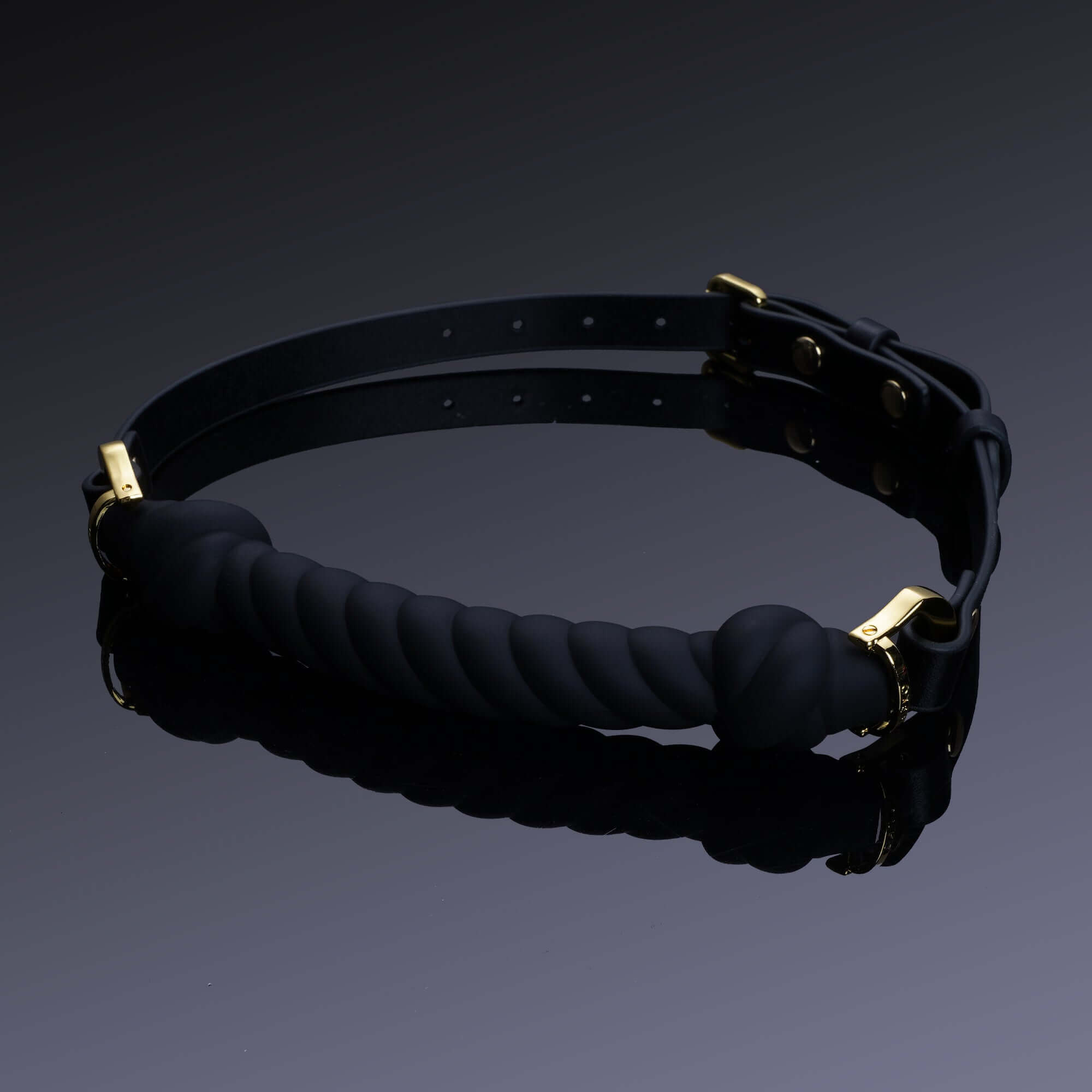 Black silicone knot-shaped mouth gag with adjustable leather strap and gold hardware on reflective surface.