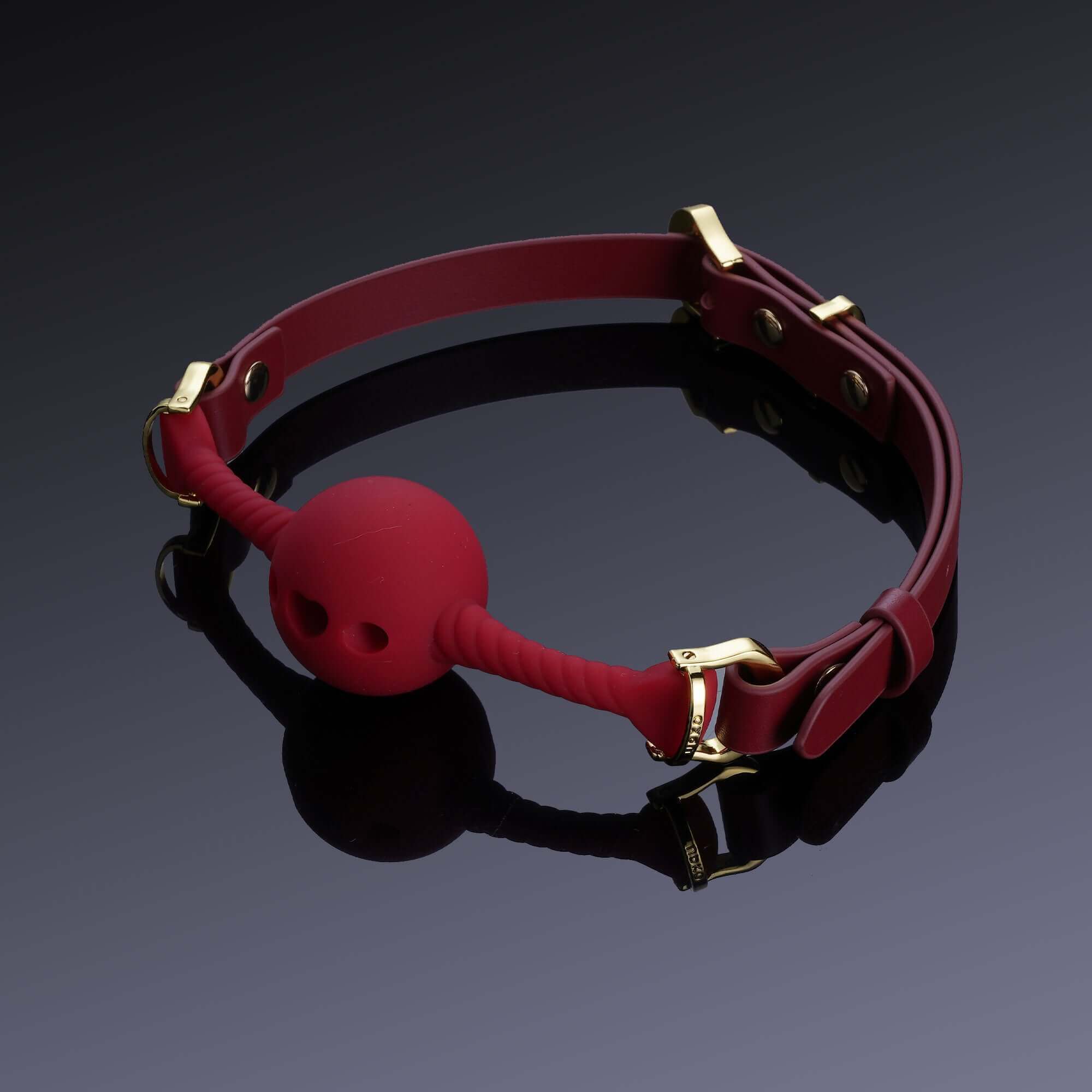 Red heart-shaped silicone mouth gag with adjustable Italian leather straps and gold accents, placed on a reflective black surface.