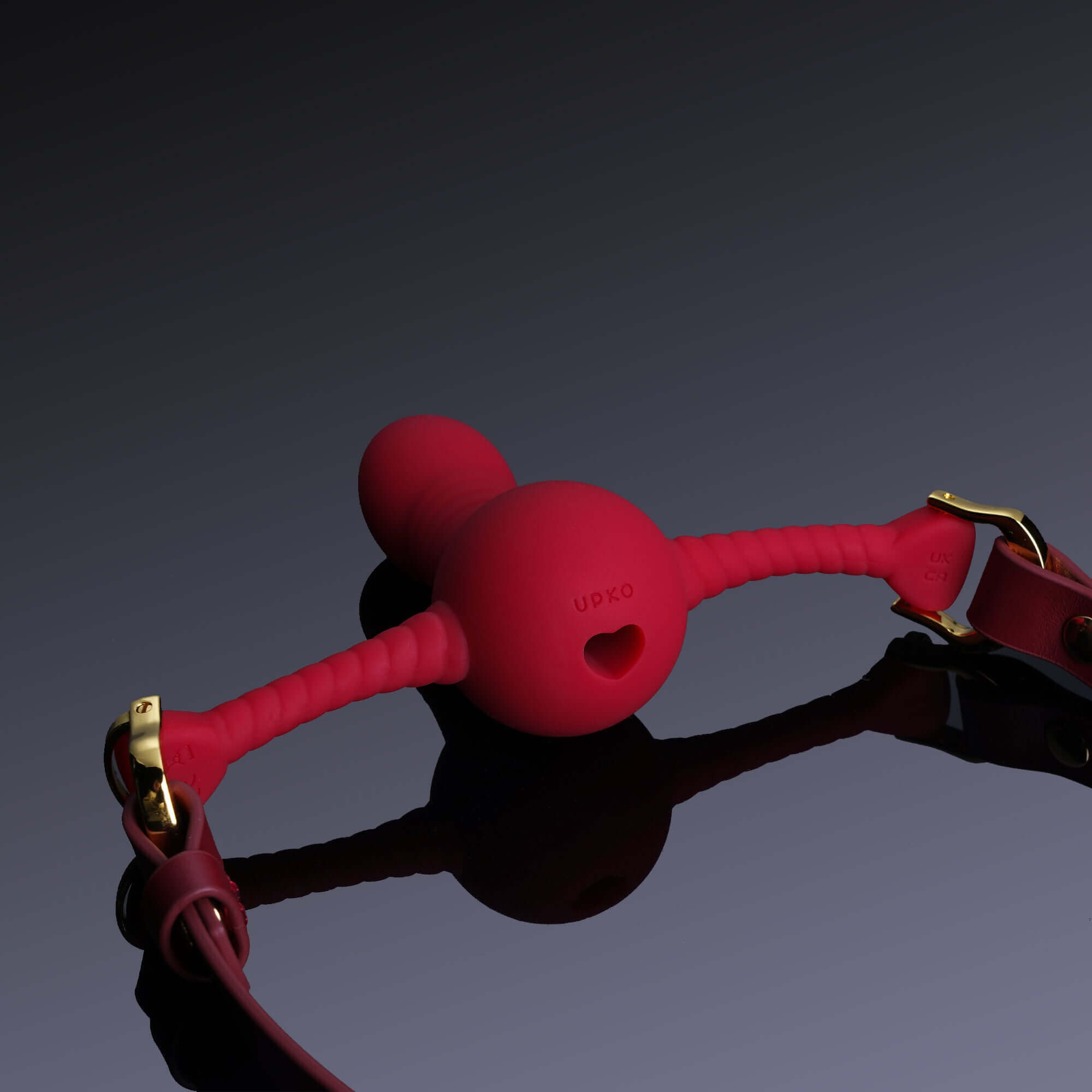 Red UPKO silicone dildo-shaped mouth gag with gold hardware and black leather straps on reflective surface.