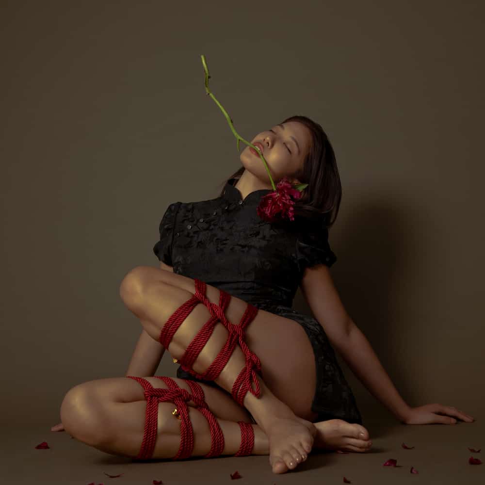 Model in black outfit adorned with red bondage rope, sitting gracefully with a flower in her mouth against a neutral background.