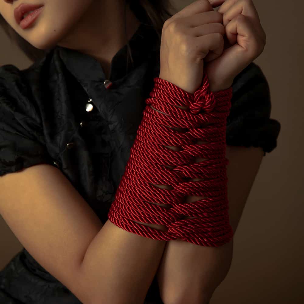 Woman's wrists bound with luxurious red Shibari bondage rope, showcasing intricate knots and texture.