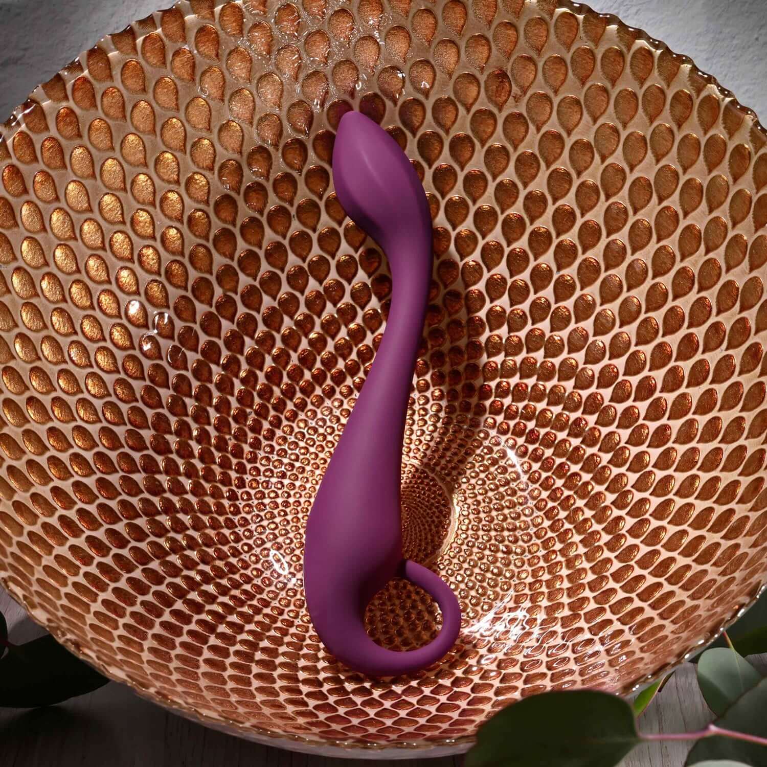 Burgundy Lochness G-spot vibrator elegantly resting in a textured bowl, showcasing its smooth design and flexible shape.